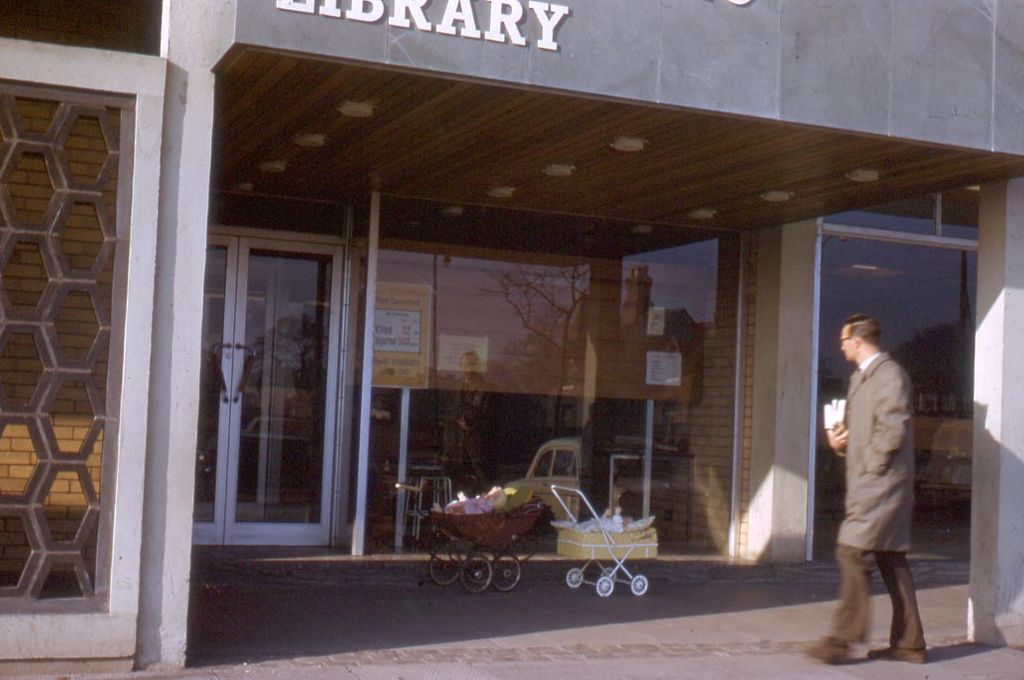 Colour photo of the glazed entrance of Kingstanding Library under a flat canopy with “LIBRARY” in large letters, a honeycomb-pattern screen wall on the left, two prams parked by the door and a man in a raincoat walking past.
