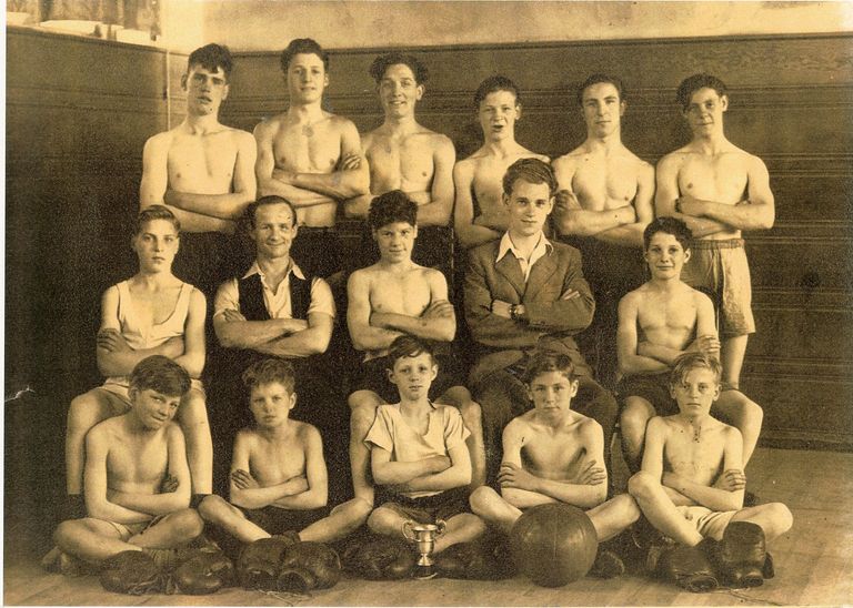 Sepia group photograph of a boys’ and young men’s boxing team in three rows in a wood-panelled hall, most bare-chested with arms folded, two men in waistcoat and jacket in the middle row, and boxing gloves, a football and a small trophy cup on the floor in front.