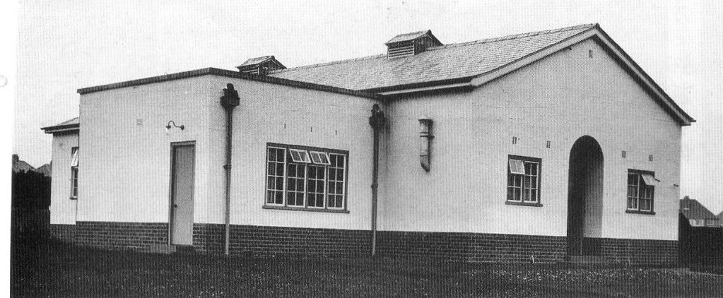 A single-storey rendered building with a dark brick base, a pitched slate roof with two small ventilator turrets, multi-paned windows, a plain side door and a round-arched porch on the gable end, standing on rough grass with houses behind.
