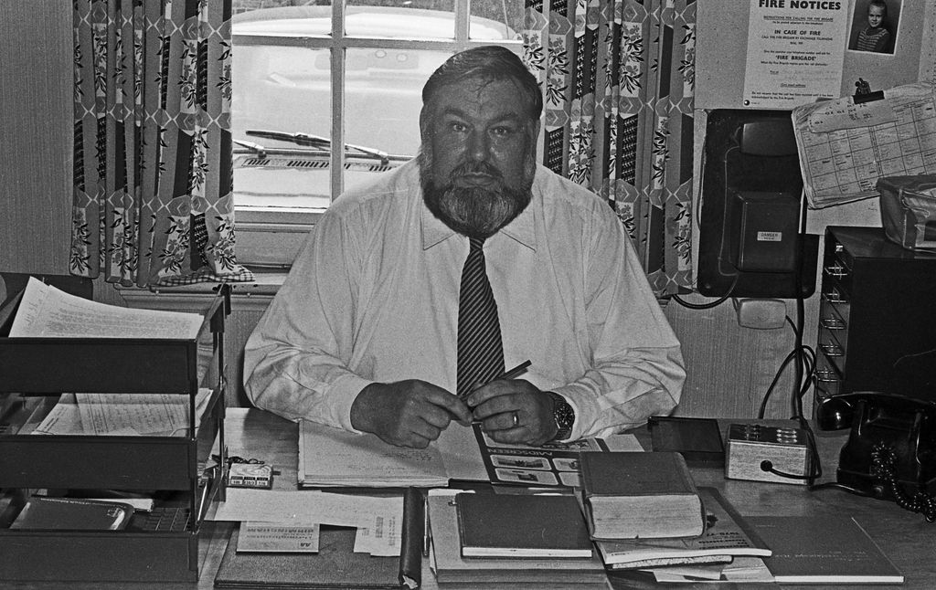 Black-and-white portrait of a large bearded man in a white shirt and striped tie seated at an office desk covered with papers and books, with patterned curtains behind him, a wall telephone and a “Fire Notices” sheet on the wall.