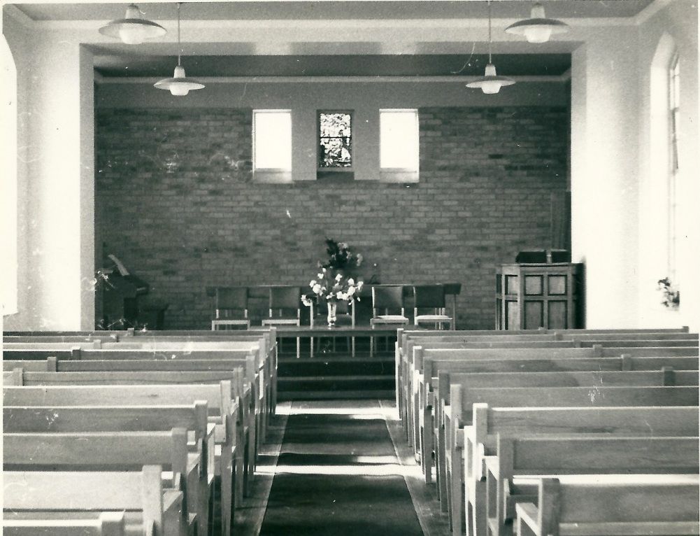 Black-and-white photo of a plain modern chapel interior: rows of light wooden pews either side of a carpeted aisle lead to a raised platform with chairs and a vase of flowers, a brick end wall with three small windows, and a pulpit on the right.