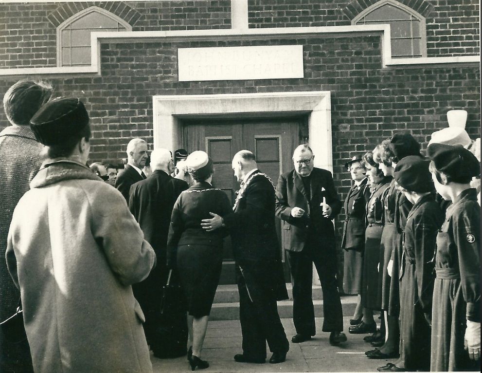 Black-and-white photo of a crowd at the door of a brick chapel inscribed “John Bunyan Baptist Chapel”: a man wearing a chain of office and a woman in a hat stand with a minister, watched by uniformed girls in berets lined up on the right.