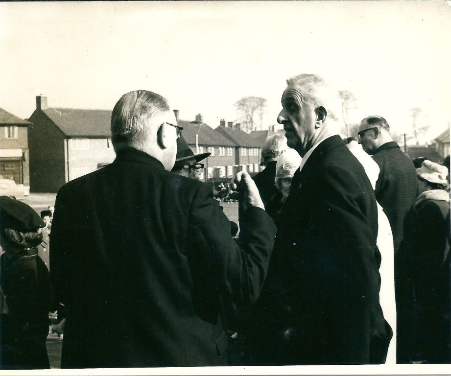 Black-and-white photo of older men in dark overcoats talking in the open air, with a woman in a hat and a uniformed girl nearby and a row of brick council houses and a green behind.
