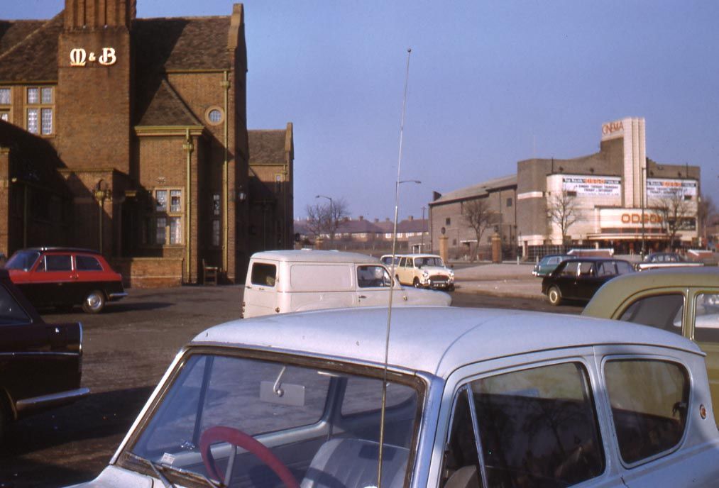 Colour photograph across a car park with 1960s cars and a van: the tall brick Kingstanding pub with an “M&B” sign on the left, and the pale Odeon with its “Cinema” fin tower and posters on the right.
