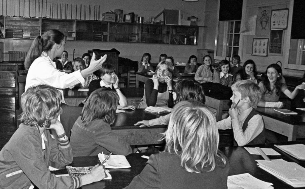 Black-and-white photo of a laughing class of girls and boys at dark laboratory benches, a woman teacher in a white lab coat gesturing as she talks; biology charts hang by the windows and shelves of books line the back wall.