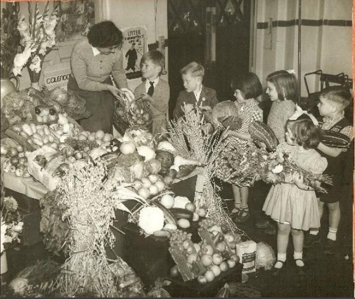 Sepia photograph of young children queuing with baskets, marrows and sheaves of corn beside a table piled with harvest produce, while a woman teacher takes a basket from a boy.