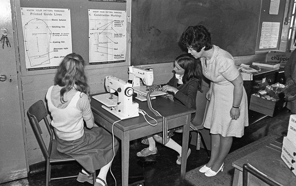 Black-and-white photo of two girls at electric sewing machines on a small table, a teacher in a light dress leaning in to watch; dressmaking pattern charts are pinned on the wall behind.