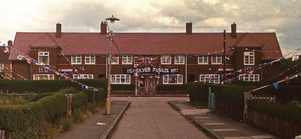 Colour photograph of a terrace of red-brick council houses hung with red, white and blue bunting and Union Jacks, with a banner reading “1952 Silver Jubilee 1977” across the front and a striped lamp post in the foreground.