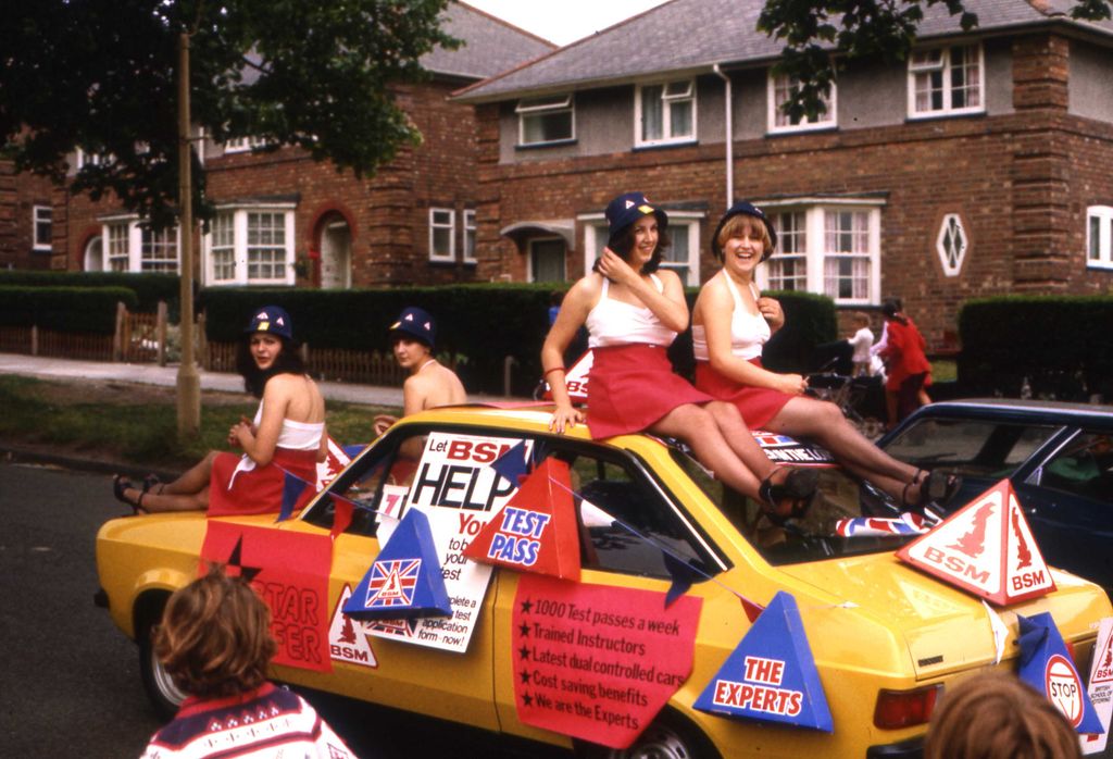 Colour photograph of four young women in red skirts, white tops and blue hats riding on a yellow car covered in British School of Motoring (BSM) posters and bunting, passing brick houses.