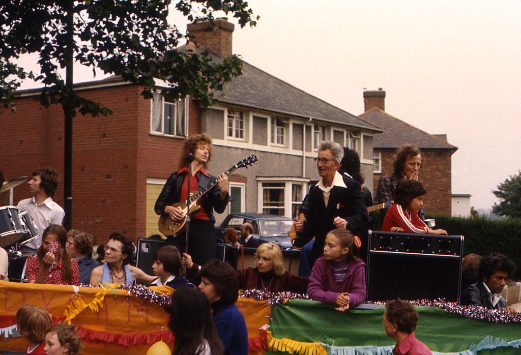Colour photograph of a decorated carnival float with a long-haired guitarist and a drummer playing among children and adults, with tinsel on the sides and pebble-dashed council houses behind.