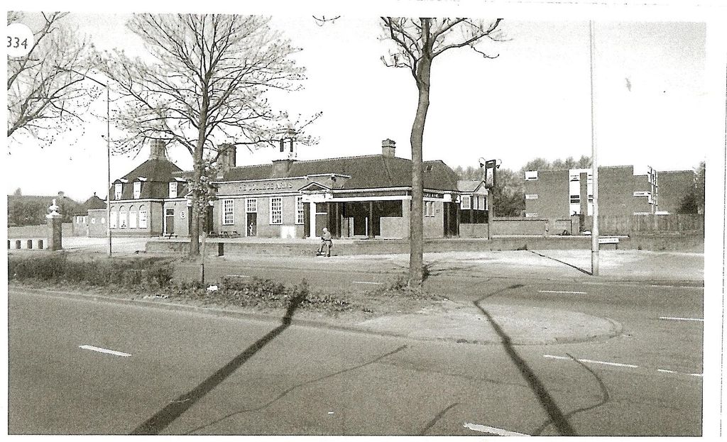 A book-plate photograph of the College Arms seen across a wide road with a planted central reservation and bare trees: a long brick pub with dormer windows, a pillared porch and a hanging sign, with flats on the right. The number “334” is printed in the top corner.