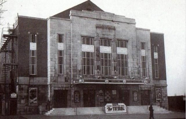 The stone-faced front of the Mayfair cinema: a tall central block with three large windows, brick side wings, a canopy lettered “Mayfair”, a steel fire escape on the left, film posters by the doors, a board on the steps and one man walking past.