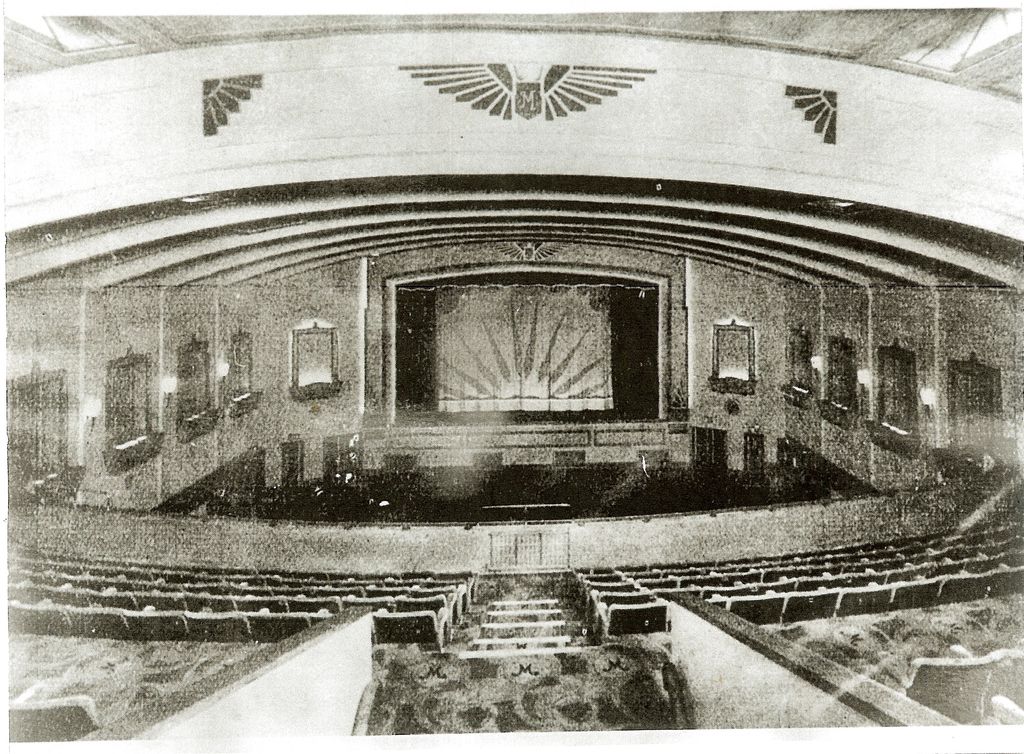 The auditorium of the Mayfair cinema seen from the back of the circle: curved balcony fronts, a large winged “M” emblem above the proscenium, a sunburst-patterned stage curtain, wall lamps in decorative frames, rows of empty seats and carpeted steps with an “M” pattern.