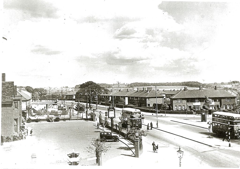 A raised view along College Road: the Crossways pub’s gate piers, hanging sign and forecourt on the left with a parked car, two double-decker buses and pedestrians on the road, rows of new brick houses across the junction, and open country and a large building on the skyline.