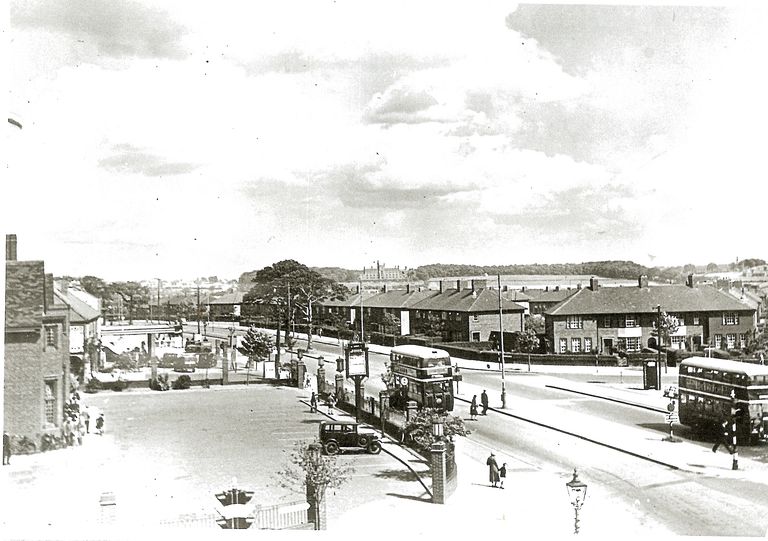 A raised view along College Road: the Crossways pub’s gate piers, hanging sign and forecourt on the left with a parked car, two double-decker buses and pedestrians on the road, rows of new brick houses across the junction, and open country and a large building on the skyline.