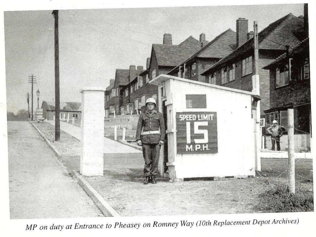 A scan of a book page: an American military policeman in a white helmet and belt stands by a white sentry box with a “Speed limit 15 m.p.h.” sign, at a gateway beside a row of houses on a sloping road; another soldier stands behind a fence. The printed caption reads “MP on duty at Entrance to Pheasey on Romney Way (10th Replacement Depot Archives)”.