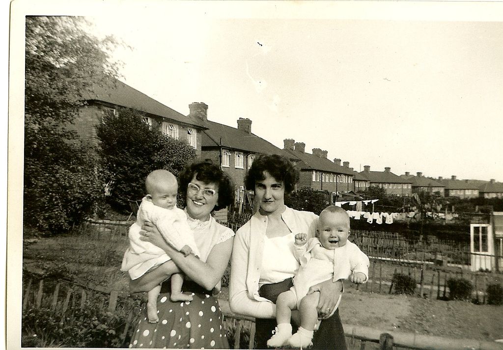 Black-and-white snapshot of two smiling young women each holding a baby in knitted clothes, standing by a garden fence; behind them a long row of brick council houses with tall chimneys, and washing on a line in the back gardens.