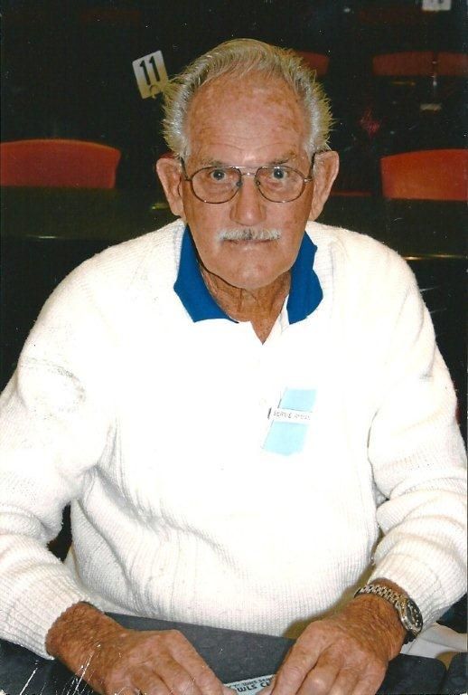 An older man with white hair, glasses and a moustache, in a white jumper with a name badge, seated at a table.