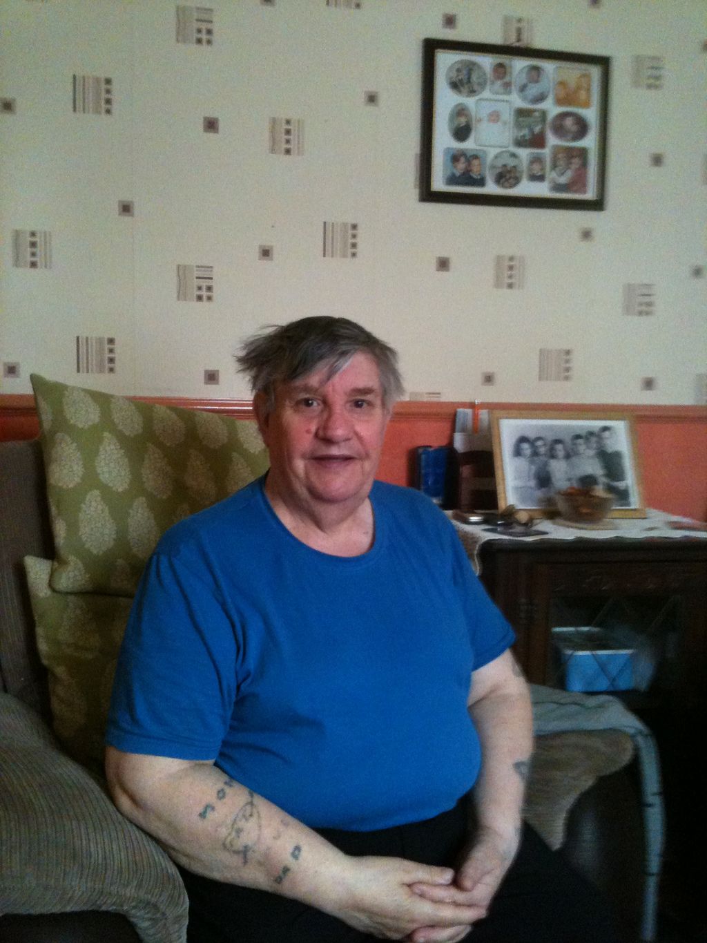 An older man with grey hair in a blue T-shirt, seated in a living room with framed family photographs.