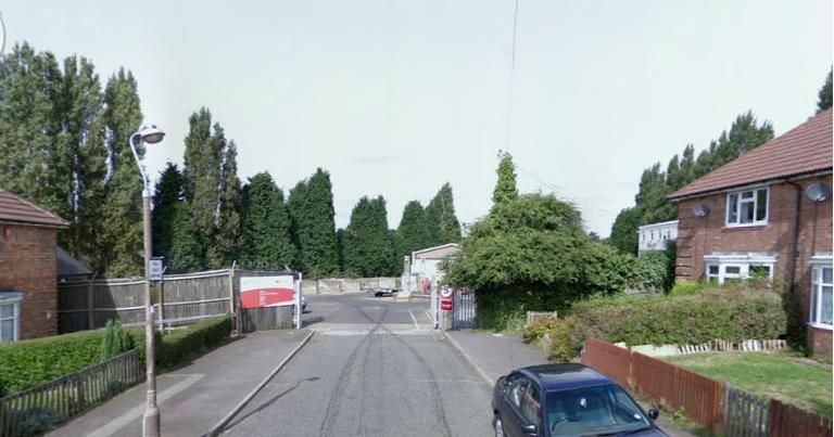 A street view along Ealing Grove to the gates of the council depot at its end, with a red sign on the fence, tall poplar trees behind, a lamp post, hedges and a car parked outside a house on the right.
