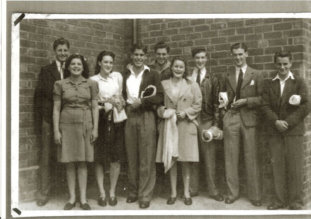 Black-and-white snapshot of nine laughing young men and women in 1940s clothes standing against a brick wall, several holding rolled towels and swimming things.