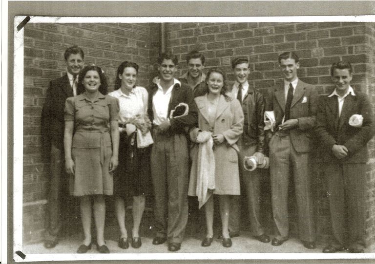 Black-and-white snapshot of nine laughing young men and women in 1940s clothes standing against a brick wall, several holding rolled towels and swimming things.