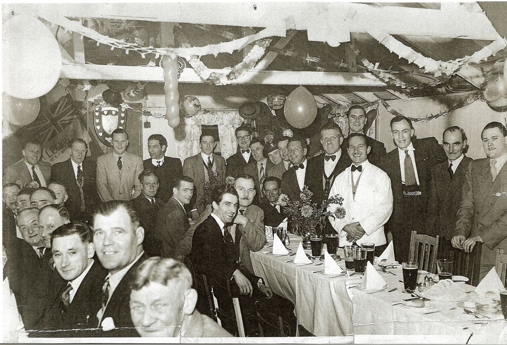 Black-and-white photograph of a crowded club dinner: rows of men in suits and bow ties at long tables laid with white cloths, pints of beer and flowers, under paper streamers and balloons; a British Legion banner and a shield hang on the wall.