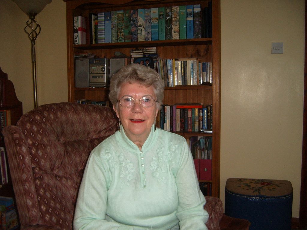 An older woman with grey hair and glasses in a pale green jumper, seated in an armchair in front of a bookcase.