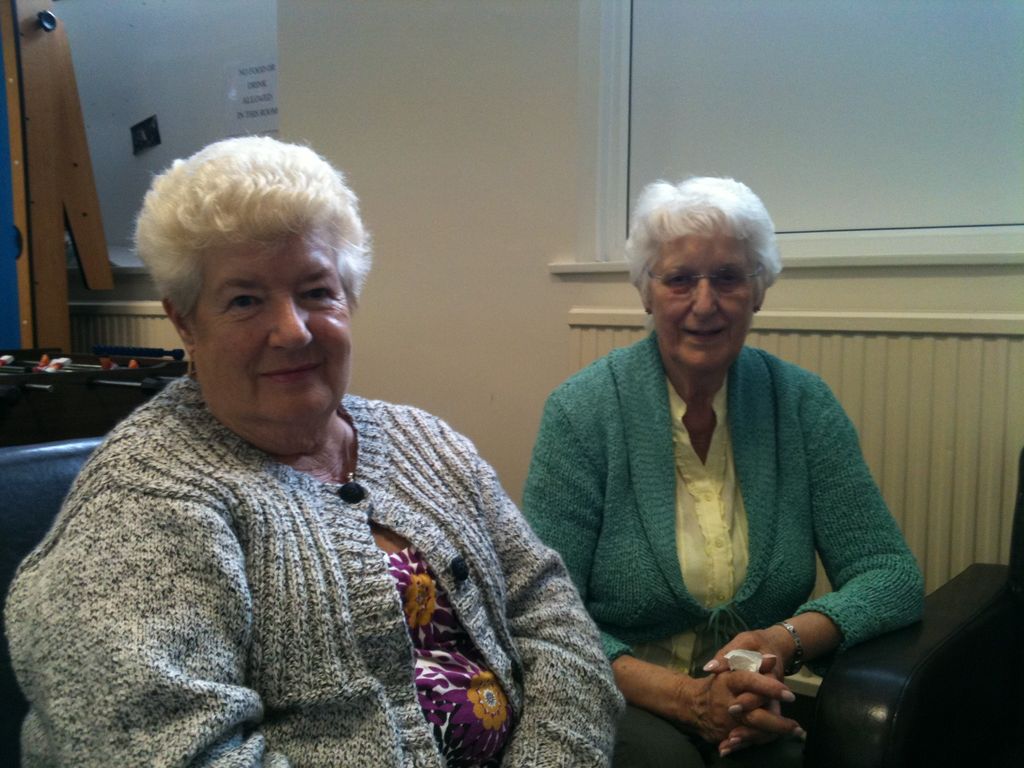 Two older women with white hair seated side by side in a community room, one in a grey knitted cardigan and one in a green cardigan.