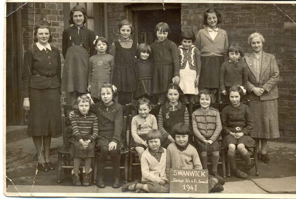 Sepia school group photograph of about twenty young girls with two women teachers outside a brick school building; two girls at the front hold a slate reading “Swanwick Junior G’s & I’s Council 1941”.
