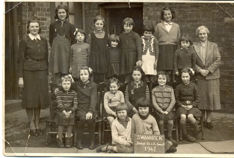 Sepia school group photograph of about twenty young girls with two women teachers outside a brick school building; two girls at the front hold a slate reading “Swanwick Junior G’s & I’s Council 1941”.