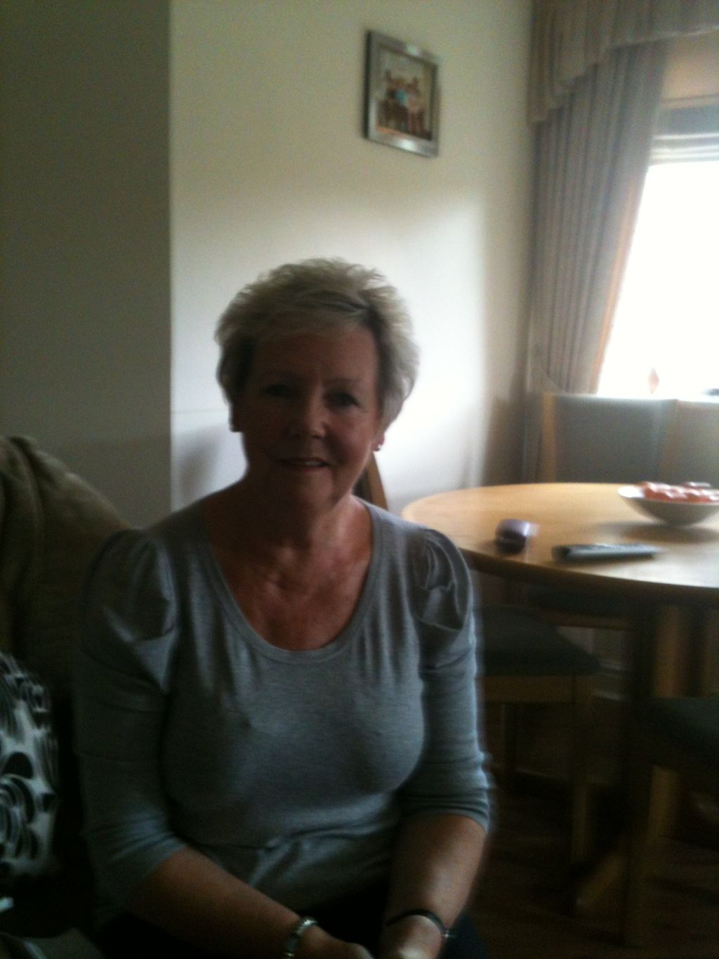 A woman with short grey hair in a grey top, seated in a living room beside a round table.