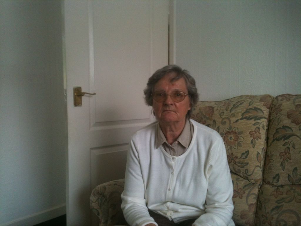 An older woman with grey hair and glasses in a white cardigan, seated on a floral sofa.