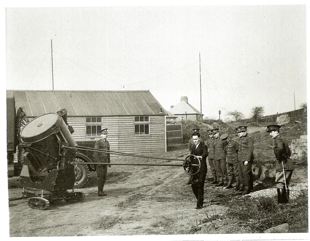 A man in a dark suit turns a control wheel linked by long rods to a large searchlight on a tracked mounting, watched by about ten soldiers in Territorial Army uniform and an officer with a swagger stick, beside a wooden hut, a lorry and a house.