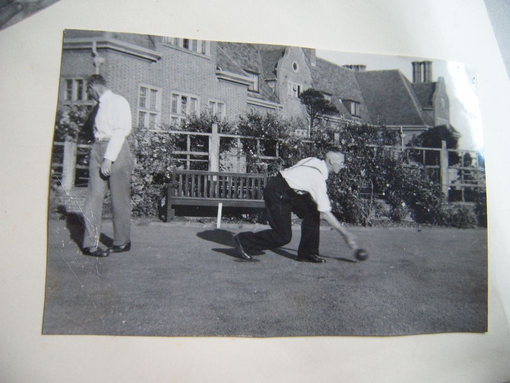 A black-and-white snapshot, mounted in an album, of an older man in braces crouching to deliver a bowl on a bowling green while a second man in a white shirt watches; a wooden bench, trellis and roses stand in front of a large gabled brick building.