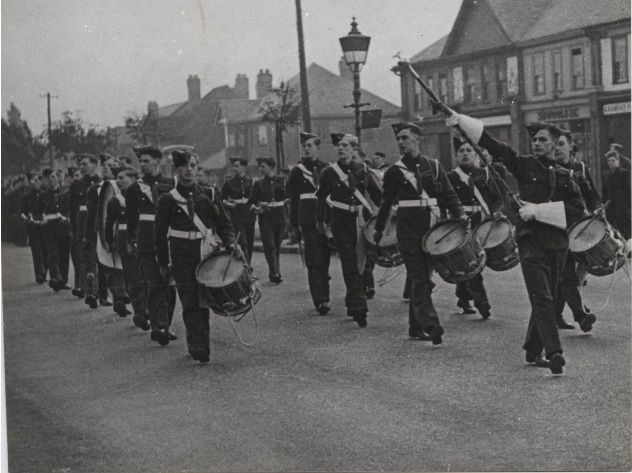 Air Training Corps cadets in forage caps and white belts march down a road behind drummers and a drum major with a raised mace; shops and a street lamp stand behind them.