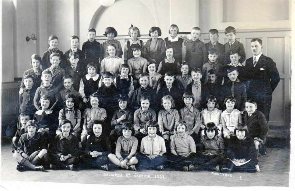 Class photograph of about forty boys and girls in four rows in a school hall, with a moustached male teacher standing at the right; written along the bottom of the print: “Dulwich Rd Junior 1932” and “Photo V. Cook”.