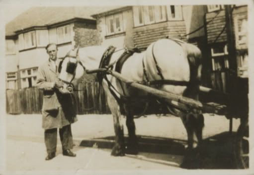 A sepia snapshot of a man in a long work coat standing at the head of a pale horse harnessed to a milk cart in a street of semi-detached houses.