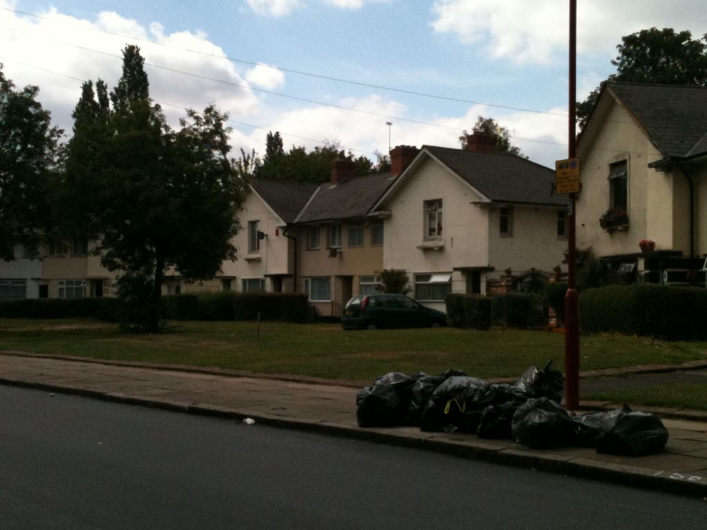 White-rendered gabled semi-detached houses behind hedges and a grass verge on Hurlingham Road, with a lamp post and a pile of black rubbish bags on the pavement in the foreground.