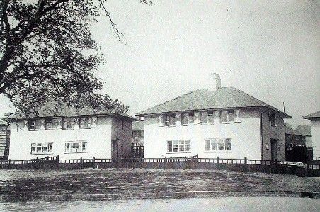 Two pairs of white-rendered semi-detached houses with hipped roofs and simple fences behind a grass verge, a large tree on the left and a street sign on the right.