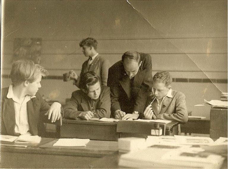 A creased sepia classroom photo of a tall teacher in a suit leaning over a desk to help a boy with his work while other teenage boys sit and stand around him; books and papers are piled in the foreground.