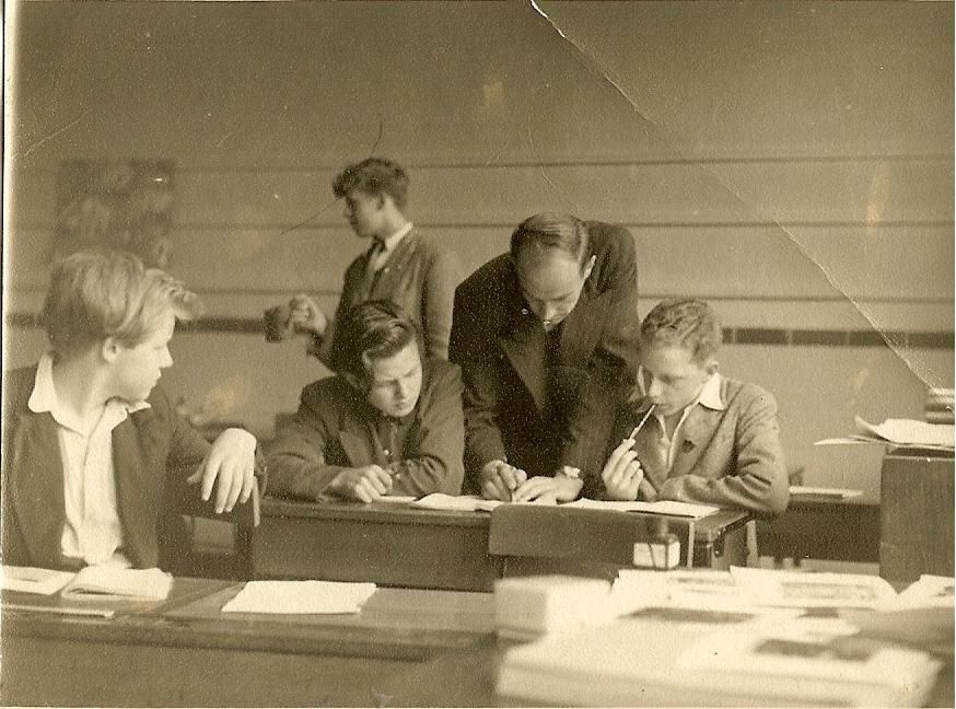 A creased sepia classroom photo of a tall teacher in a suit leaning over a desk to help a boy with his work while other teenage boys sit and stand around him; books and papers are piled in the foreground.
