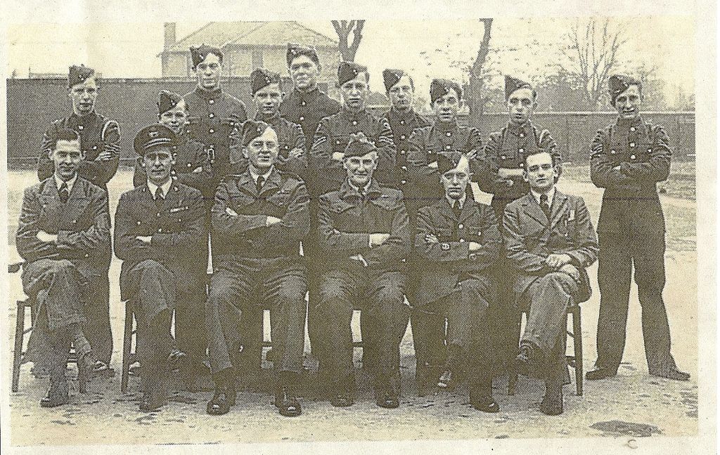 Group portrait of about seventeen Air Training Corps cadets in forage caps standing behind six seated officers and instructors, most with arms folded, in front of a brick wall and a house.
