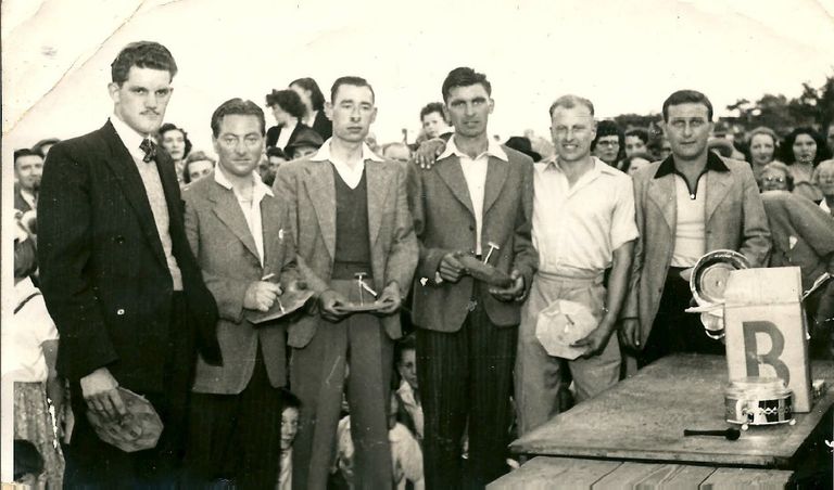 Six men in jackets and open-necked shirts stand in a row holding small cake stands, with a crowd behind them and a prize table with a lettered “B” board and trophies at the right.
