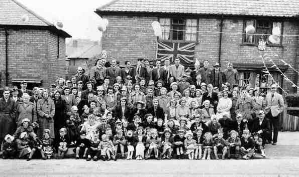 A large group of residents – several rows of adults and children seated on the pavement in front – gathered in the street for the coronation, with a Union flag on a house, bunting and balloons.