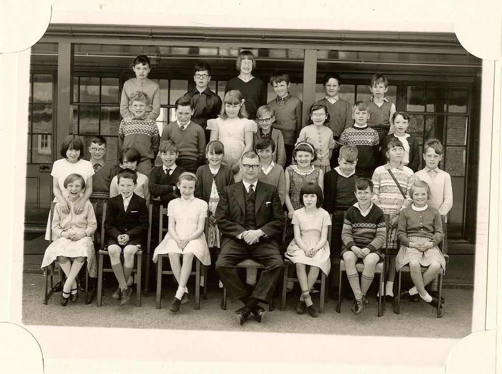 Class of about thirty junior-school children in four rows on the playground, a male teacher in glasses and a three-piece suit seated in the centre of the front row, in front of a veranda with glazed doors.