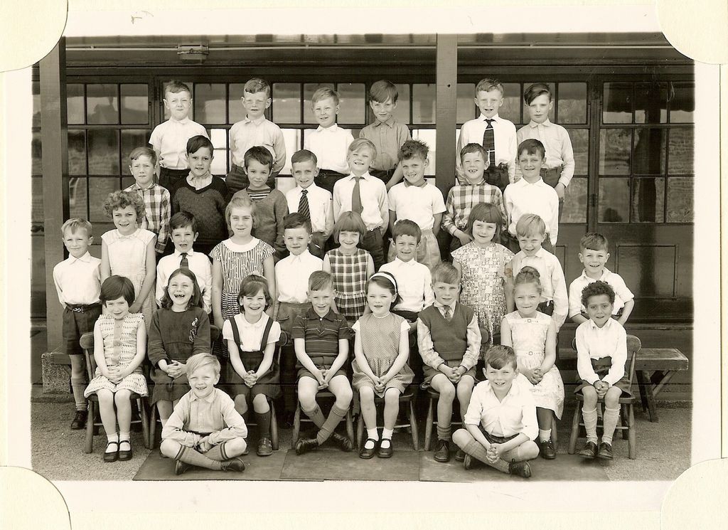 Class of about forty infants in five rows, two boys sitting cross-legged on a mat in front, in front of a veranda with glazed doors; no teacher appears.