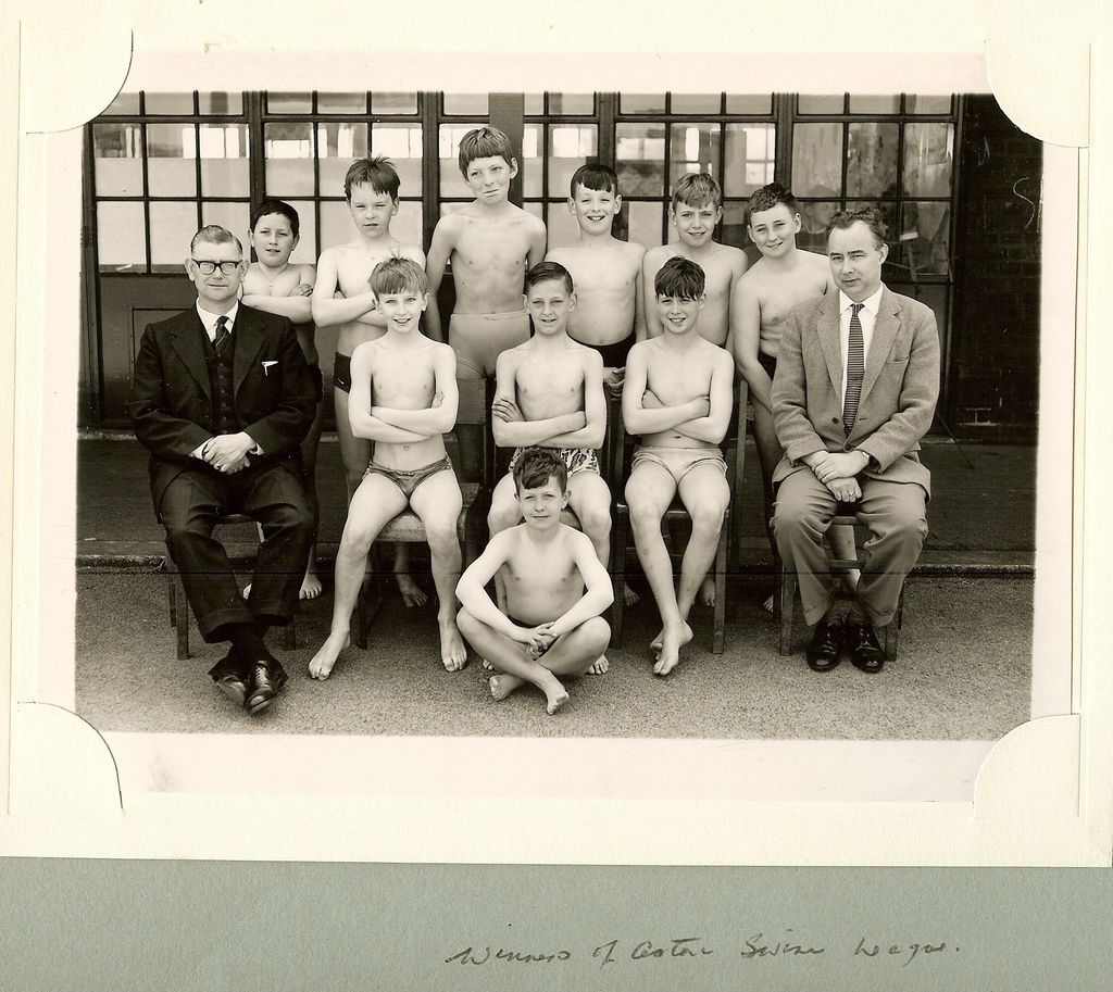 A school team photograph of eleven boys in swimming trunks with two male teachers in suits, in front of the school’s glazed doors, mounted in an album with the handwritten caption “Winners of Aston Swim League”.