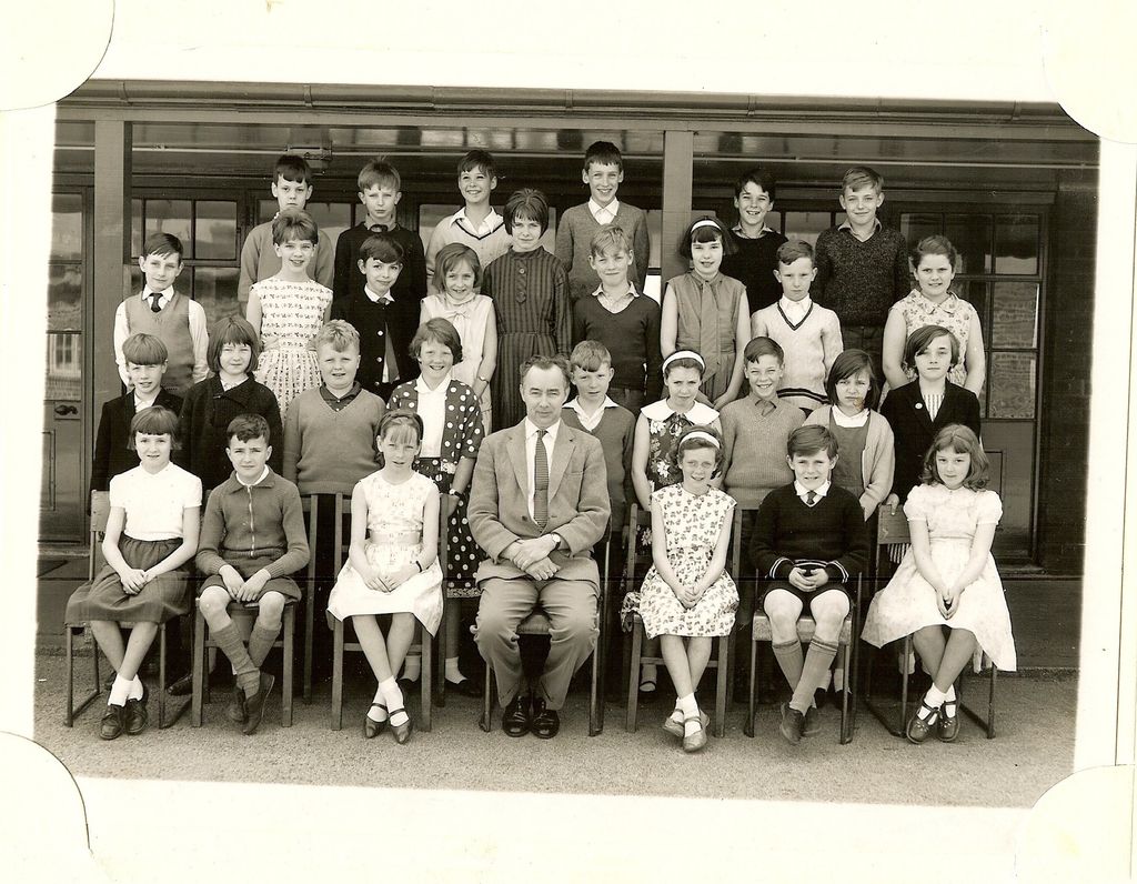 Class of about thirty-three junior-school children in four rows, a male teacher in a light jacket and striped tie seated in the centre of the front row, in front of glazed classroom doors.
