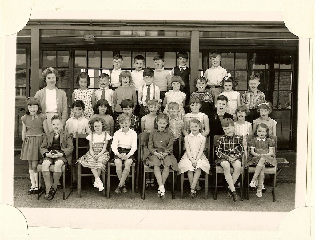 Class of about thirty-four young children in four rows, a smiling woman teacher in a cardigan standing at the left, in front of a veranda with glazed doors.